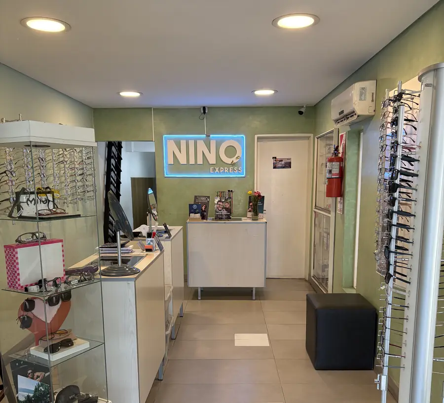 El local por dentro: los exhibidores de anteojos a los costados y, al fondo, el cartel de Nino Express iluminado en azul sobre la pared verde